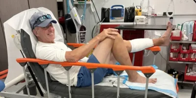 John Schweizer sentado en una cama de hospital tras la mordedura de tiburón en Bahamas, con la parte inferior de la pierna izquierda vendada