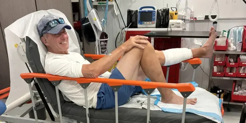 John Schweizer sentado en una cama de hospital tras la mordedura de tiburón en Bahamas, con la parte inferior de la pierna izquierda vendada
